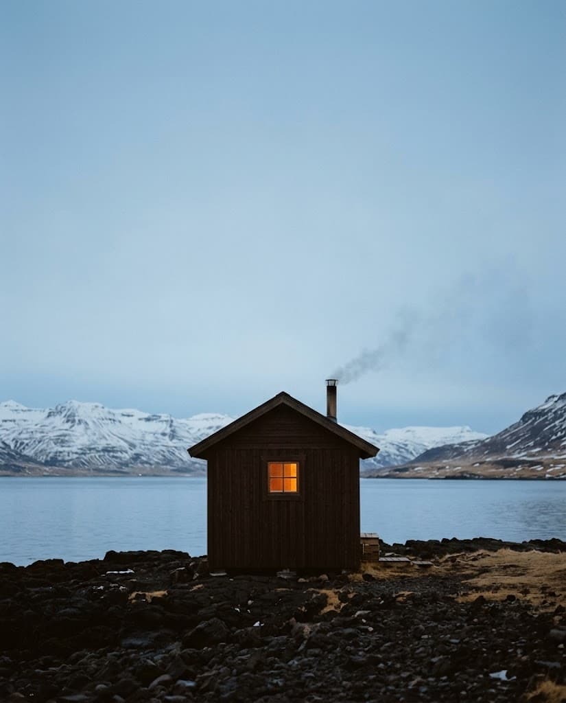 A small dark timber sauna with a glowing window on an Icelandic fjord shore, snowy mountains behind